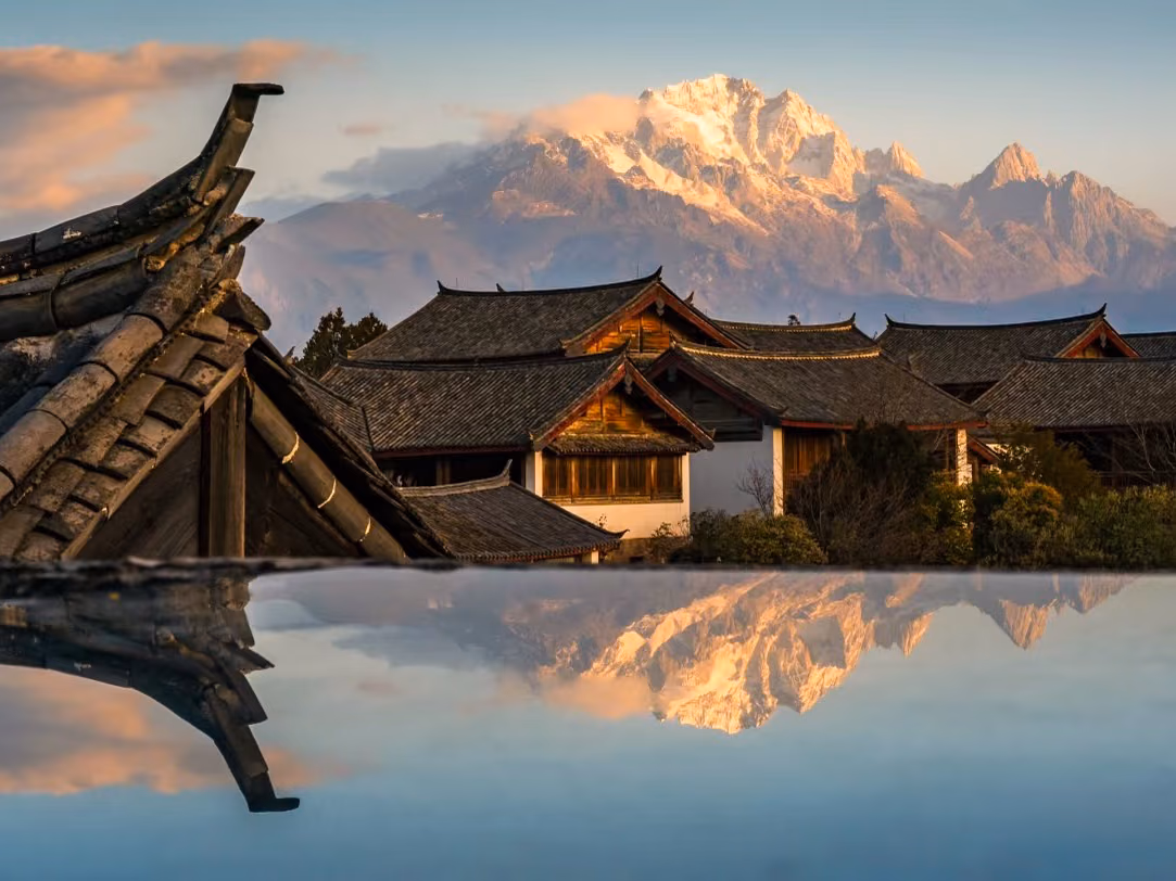 Naxi rooftops with Jade Dragon Snow Mountain glowing at sunset, reflected in water, Yunnan