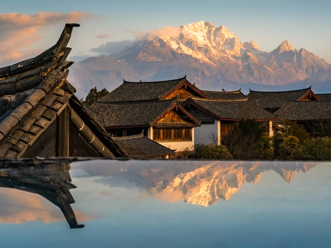 Naxi rooftops with Jade Dragon Snow Mountain glowing at sunset, reflected in water, Yunnan