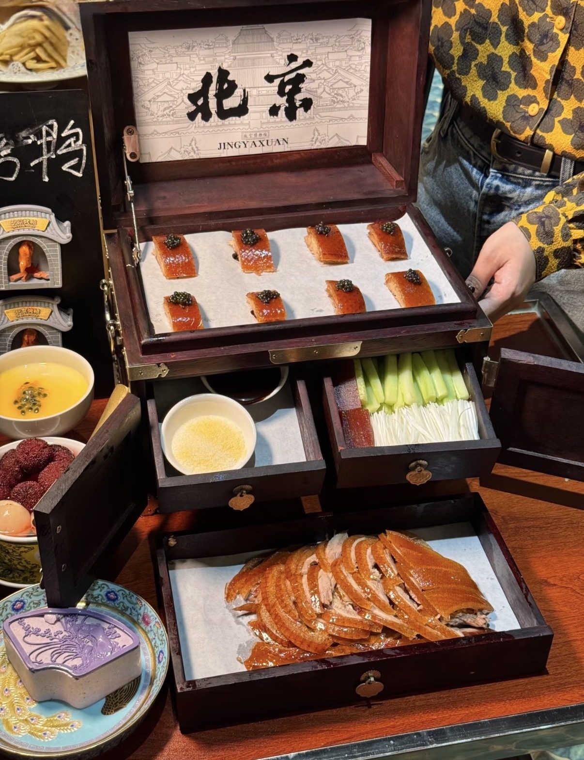 Lacquered slices of Peking duck served in a tiered wooden presentation box, condiments in its drawers