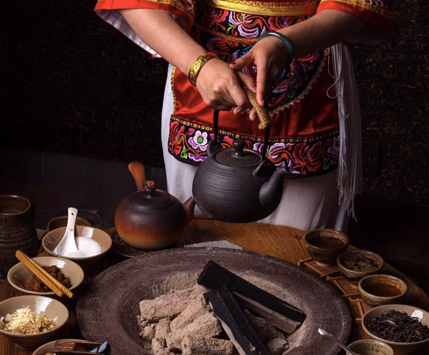A tea master warming tea over a charcoal hearth, surrounded by small bowls of leaves and spices