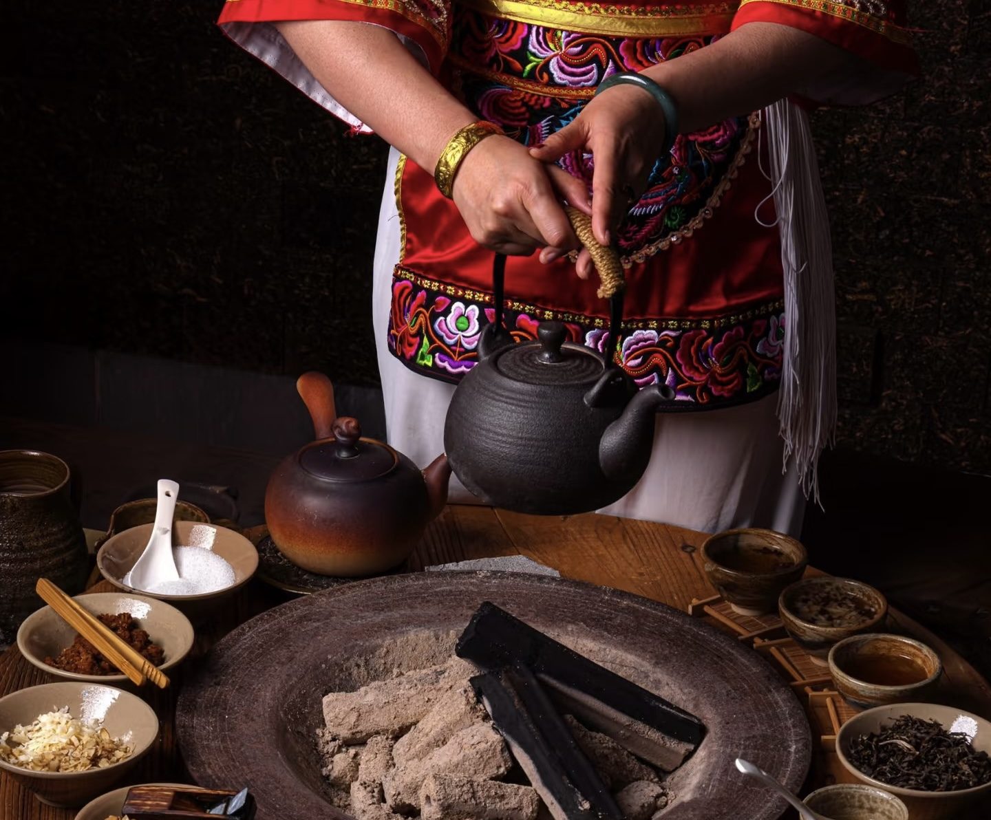 A tea master in embroidered dress warming tea over a charcoal hearth, surrounded by small bowls of leaves and spices