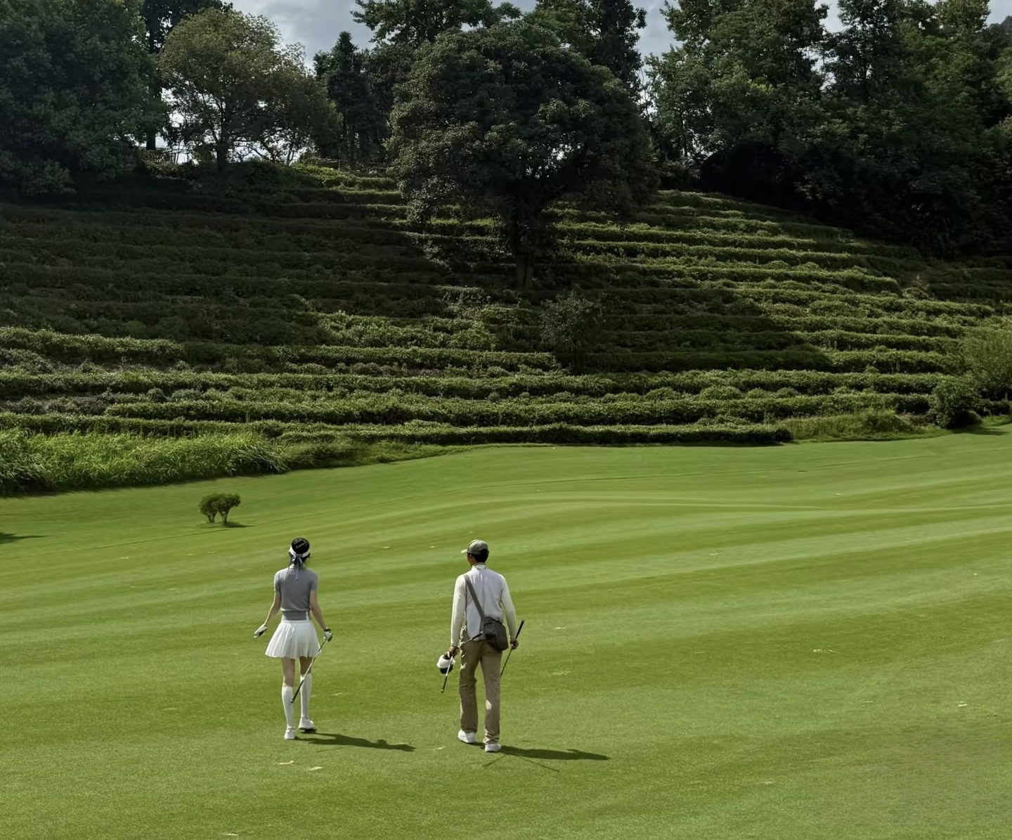 Golfers walking a Fuchun fairway beneath terraced walls of Longjing tea