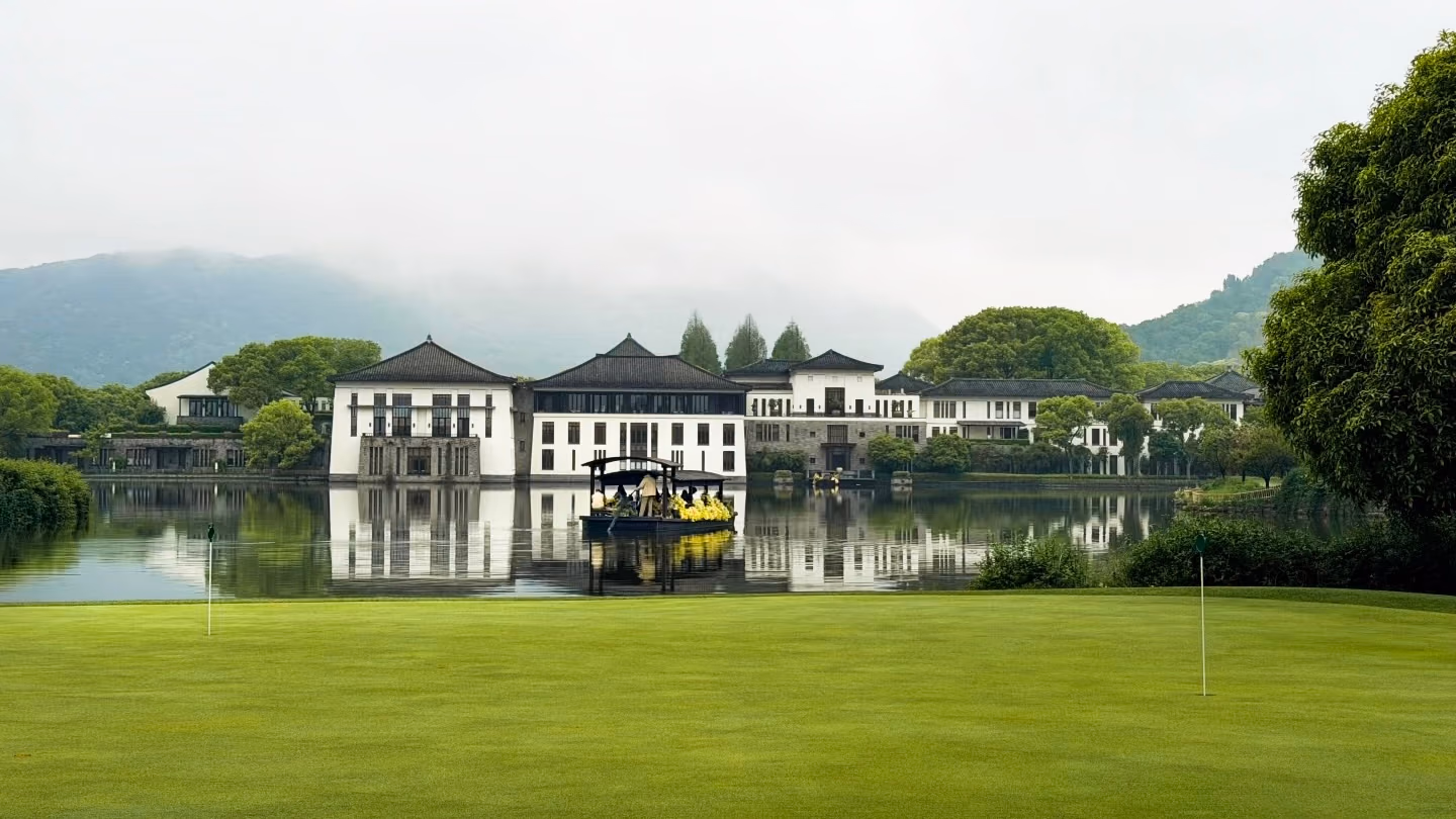 A golf green beside a still lake reflecting a classical Chinese resort, misty mountains behind
